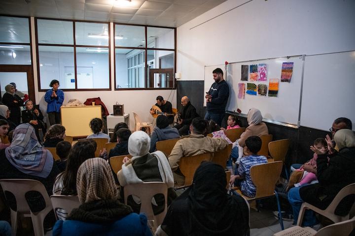 Des enfants chantent en chœur une chanson traditionnelle palestinienne. Devant deux musiciens venus pour eux. Derrière eux, les parents se regardent en riant