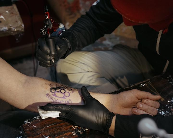 Au fond du bar, un homme en cagoule rouge et gants noirs tatoue au choix : un poing américain entouré du texte « Welcome to Ménilmontant » ou un ange, emblèmes du bar le Saint Sauveur