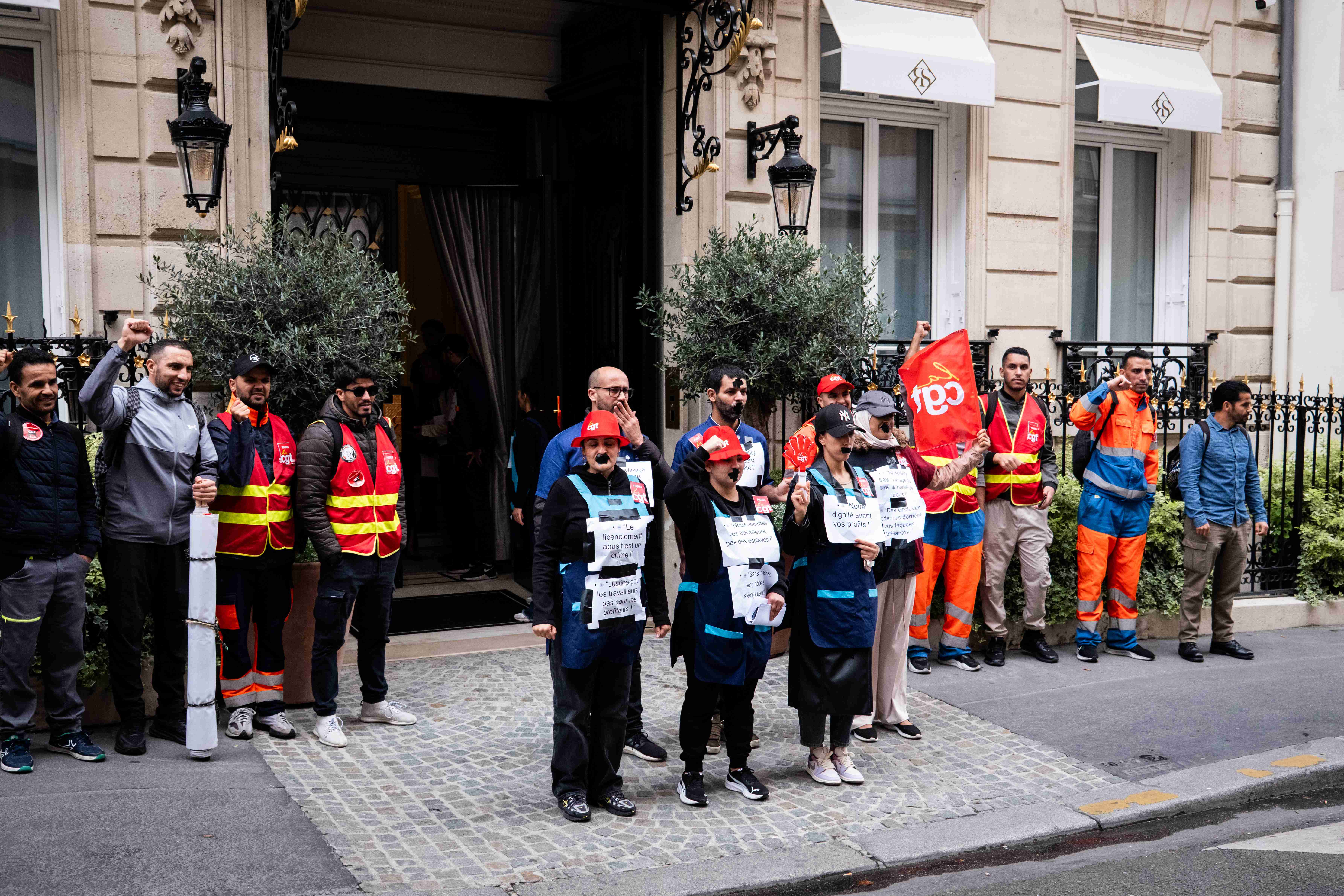 Poings levés et scotch noir sur la bouche, huit femmes et valets de chambre sans papiers, se tiennent devant la porte cochère de la luxueuse « Maison Bauchart »